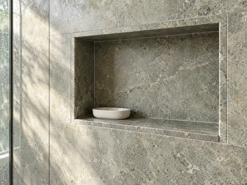 A recessed grey-green stone shower niche has supported side returns and a subtly sloped shelf.