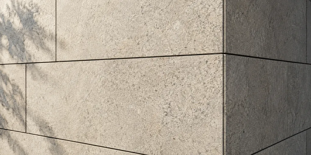 Sunlight grazes warm grey stone panels and narrow joints around an exterior corner.