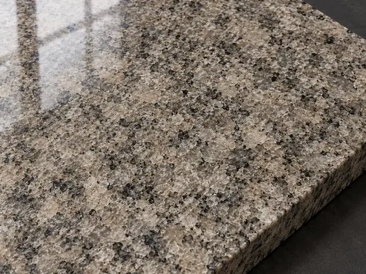Polished warm-grey granite face with interlocking mineral grains and a soft window reflection.